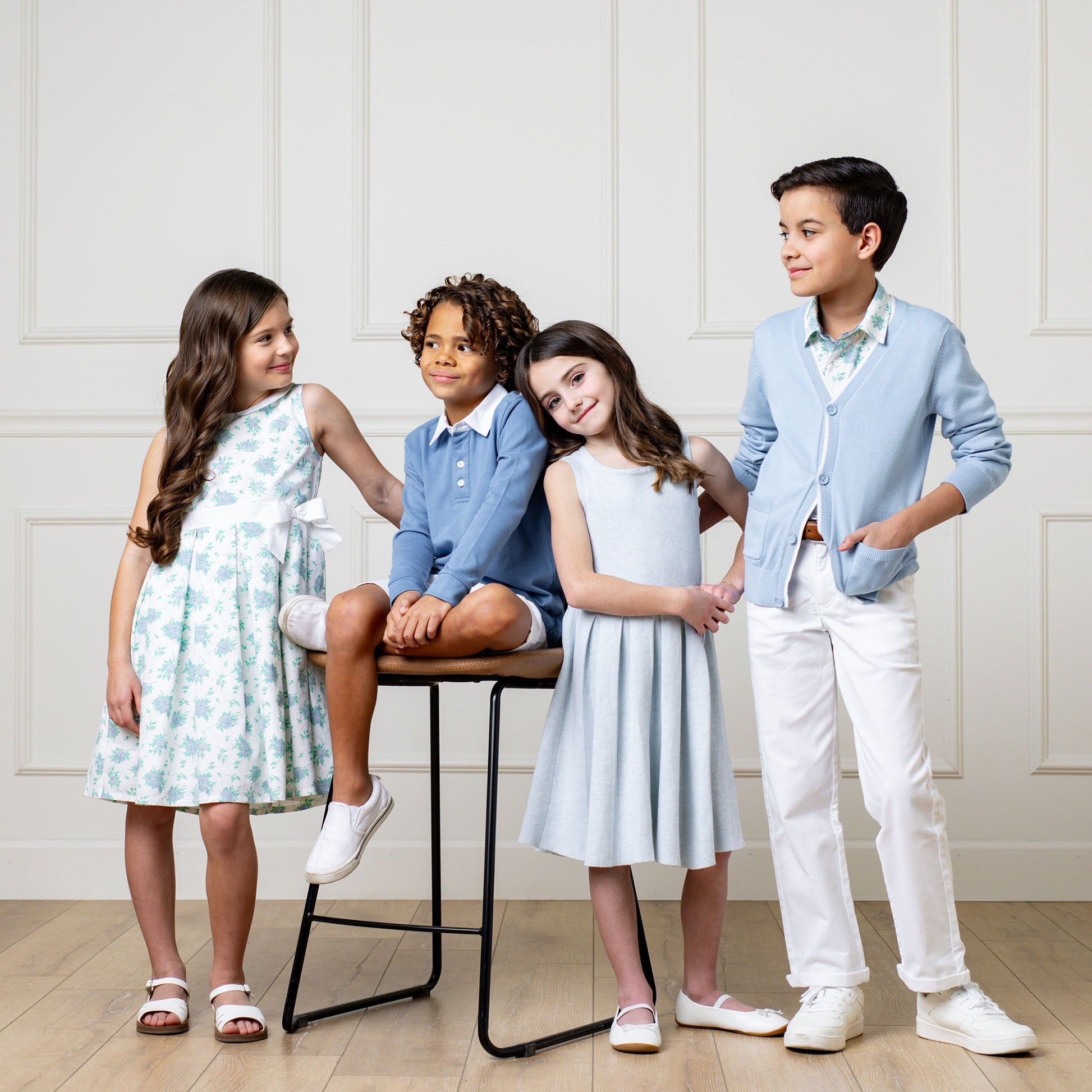 Four children posing together in a room with light-colored walls and wooden floor.