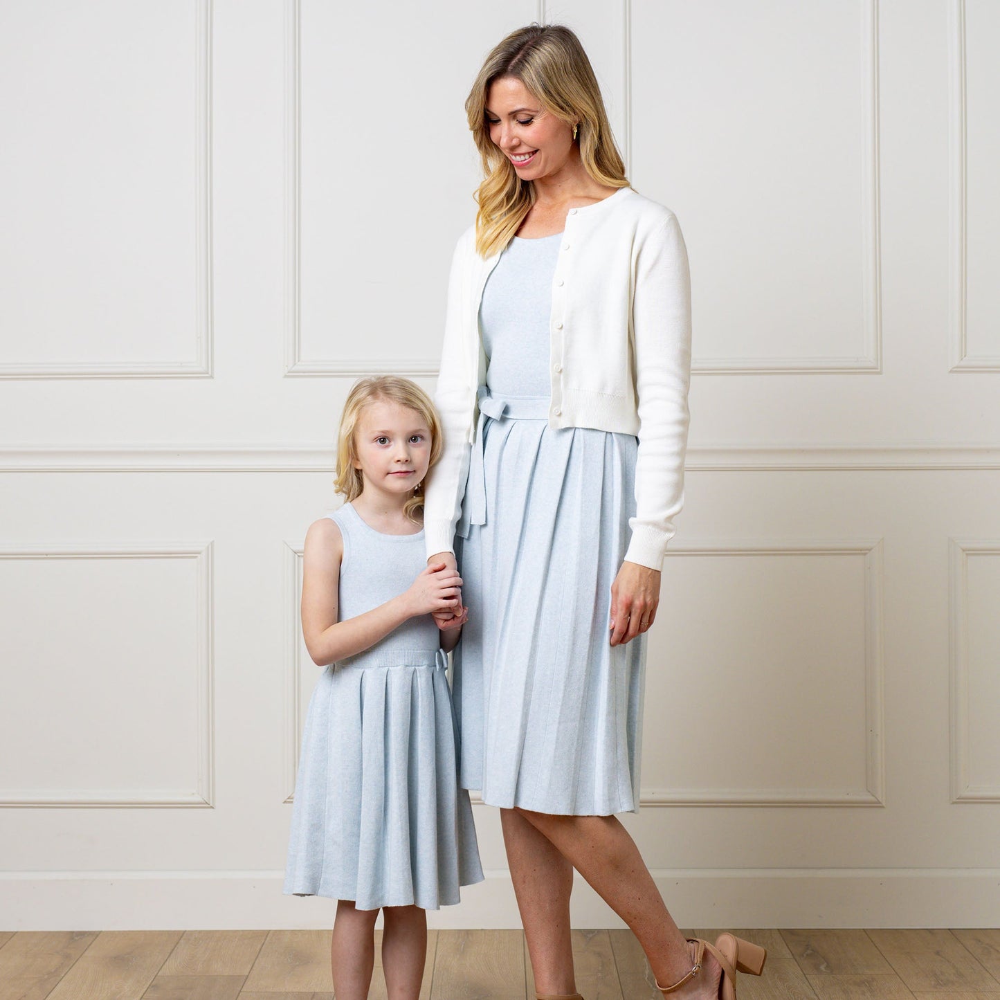Woman and young girl wearing matching light blue dresses with white cardigans against a white paneled wall.