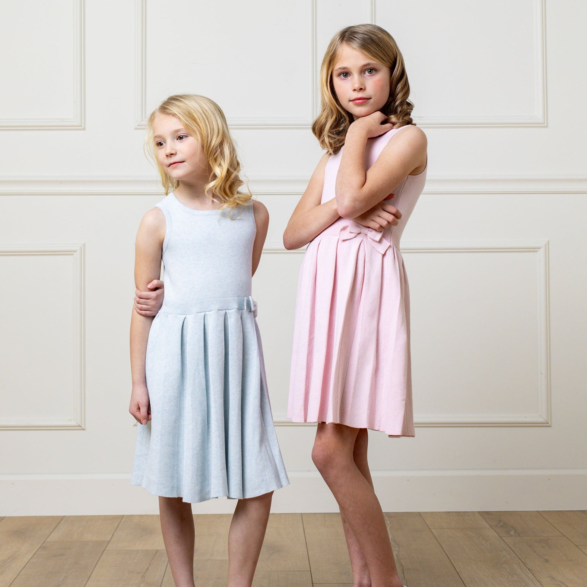 Two young girls wearing light blue and pink dresses standing against a white paneled wall.