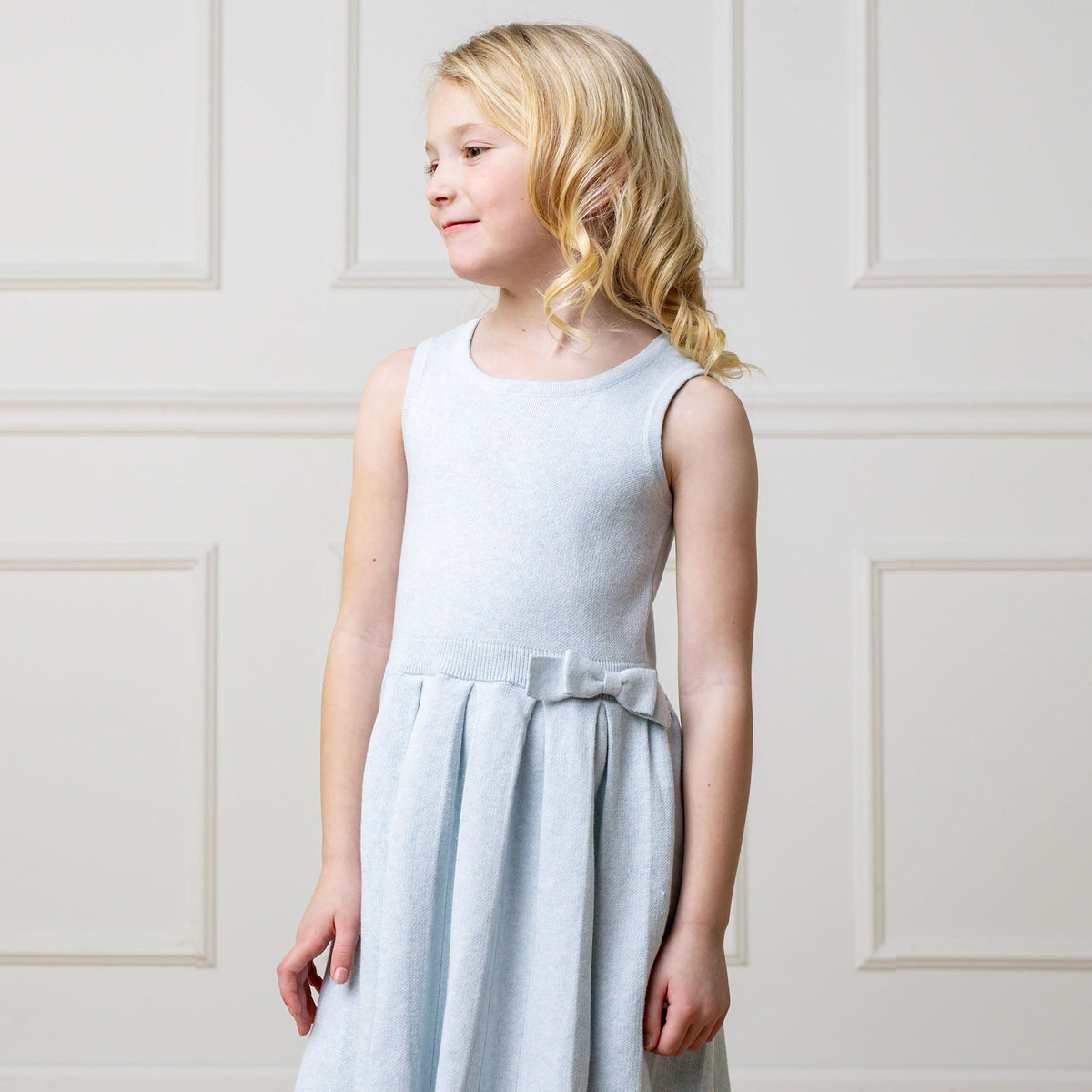 Young girl wearing a light blue dress with a bow detail against a white paneled wall.