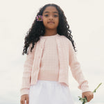 Young girl wearing a light pink cardigan over a pink top with white pants, holding a small plant.