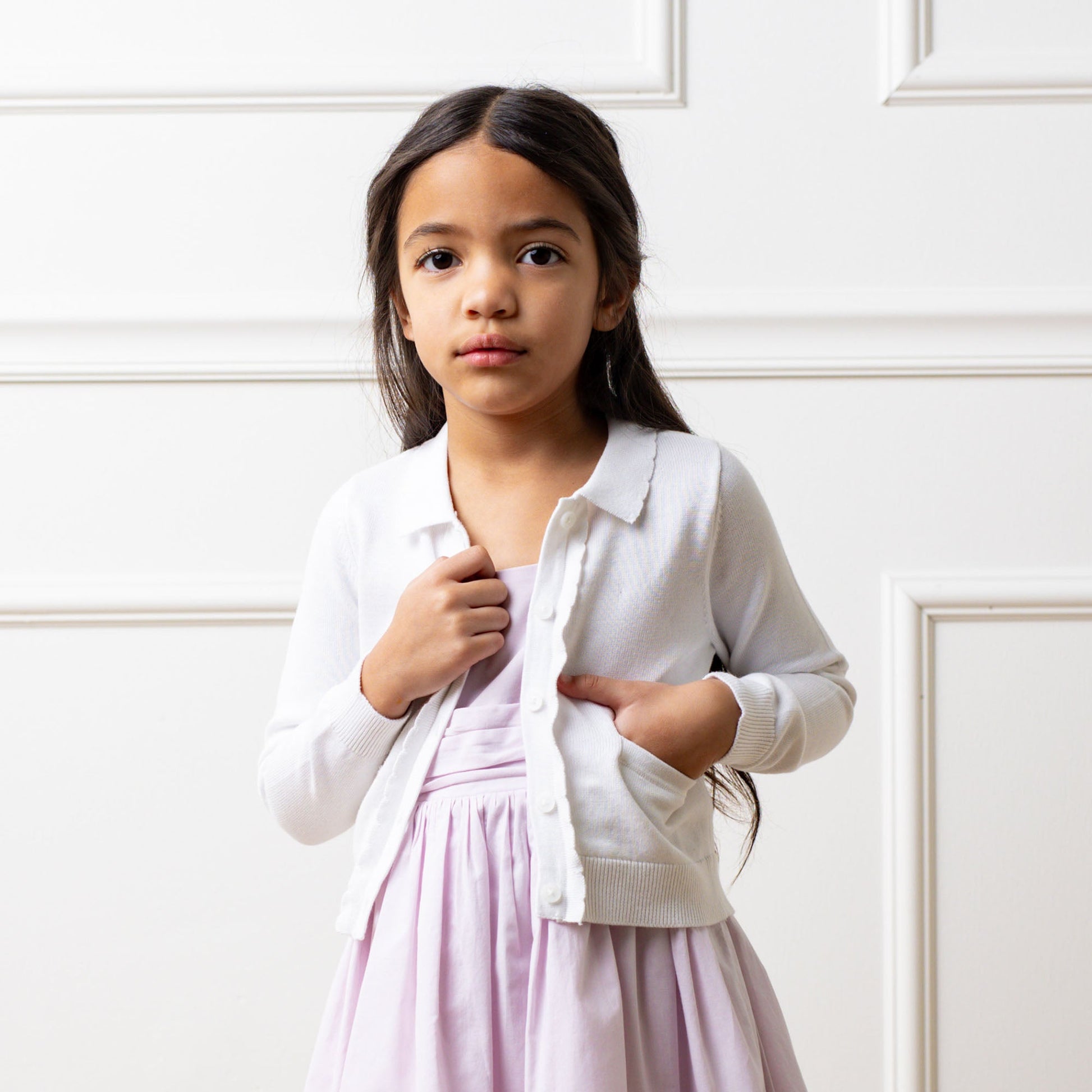 Young girl wearing a white cardigan over a pink dress against a white wall.