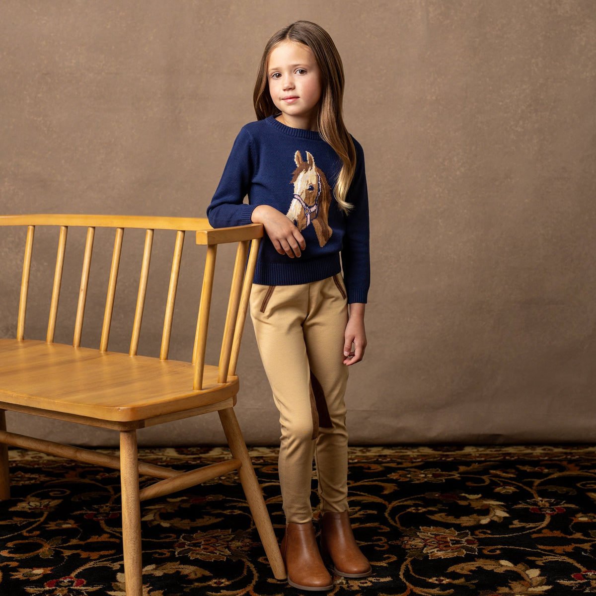Young girl in a navy sweater with a horse design, beige pants, and brown boots standing next to a wooden chair.