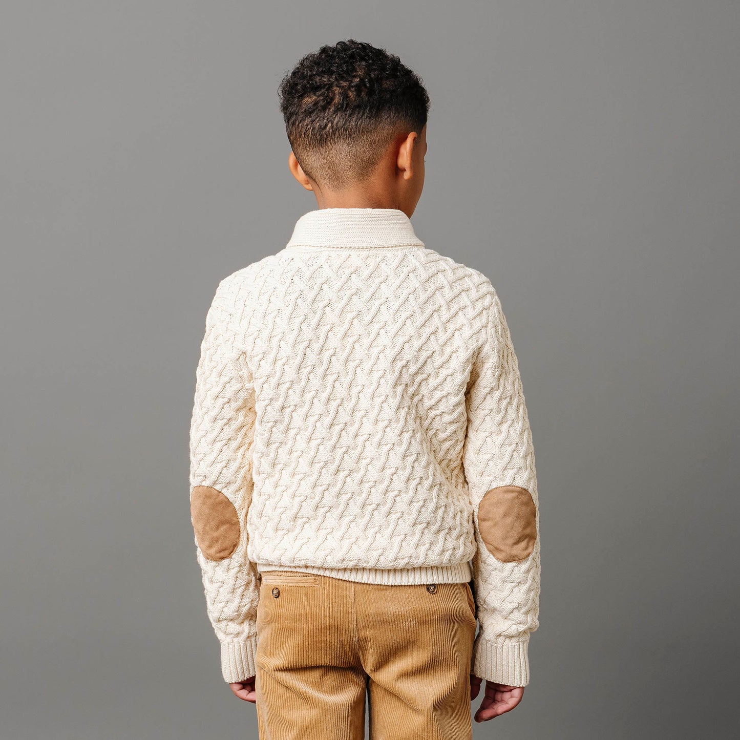 Child wearing a cream knitted sweater with brown elbow patches on a gray background
