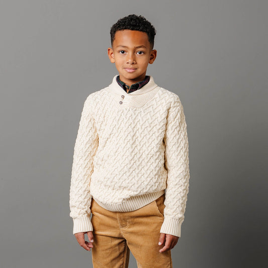Young boy wearing a cream sweater and brown pants against a gray background