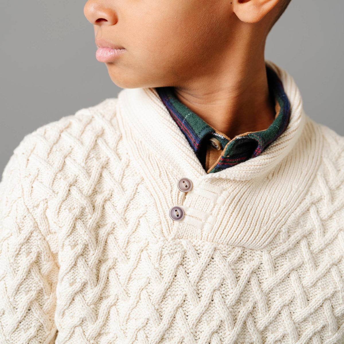 Close-up of a person wearing a cream cable knit sweater with a plaid shirt underneath.