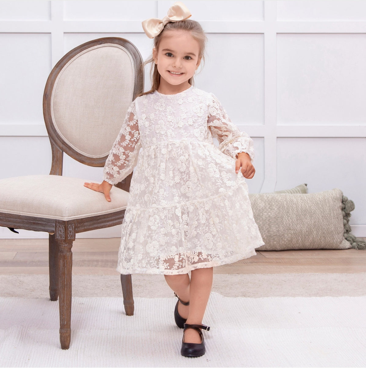 Young girl in a white lace dress standing in a room with a wooden chair and decorative pillows.