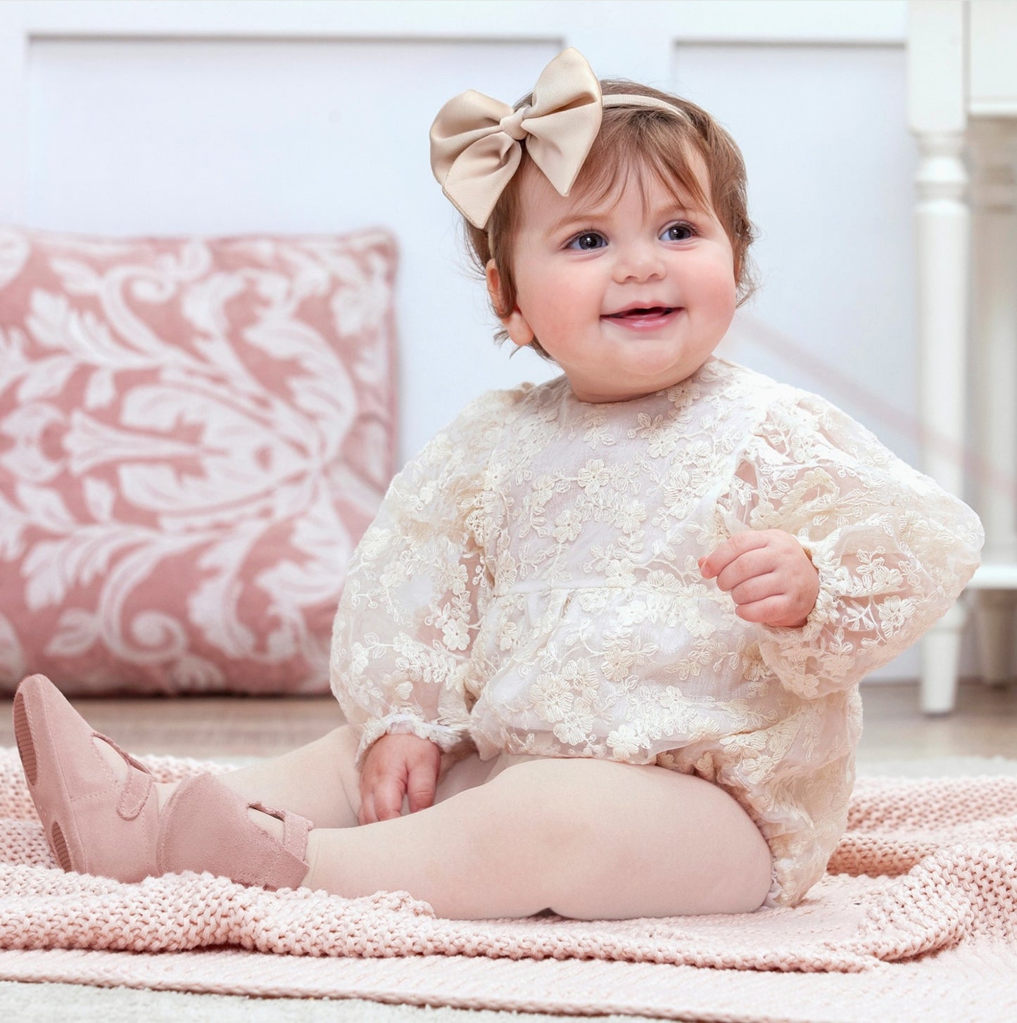 Baby sitting on a pink blanket wearing a lace romper and bow headband.