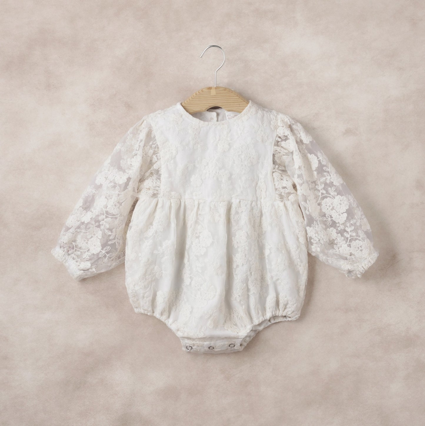 White lace baby romper on a wooden hanger against a beige background
