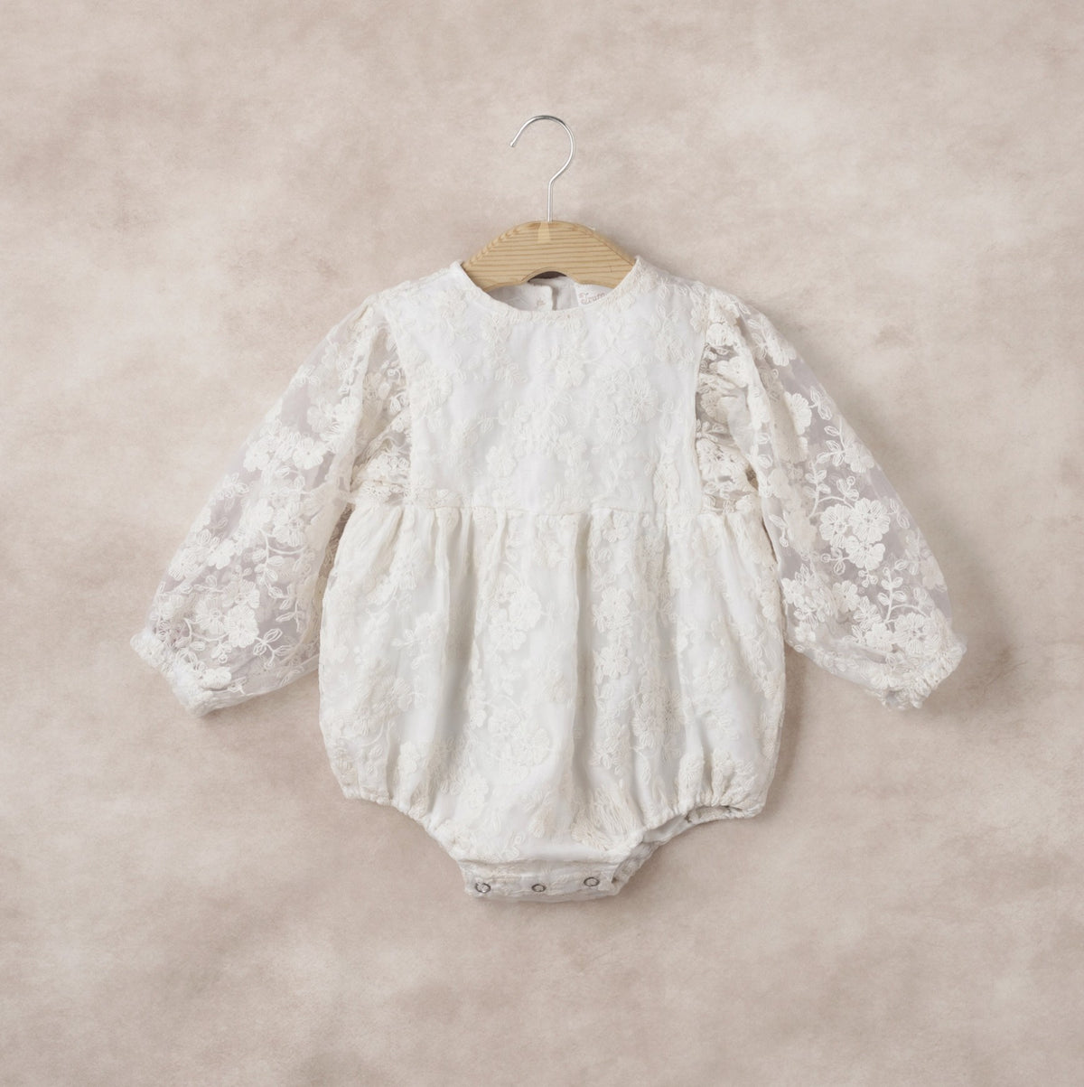 White lace baby romper on a wooden hanger against a beige background