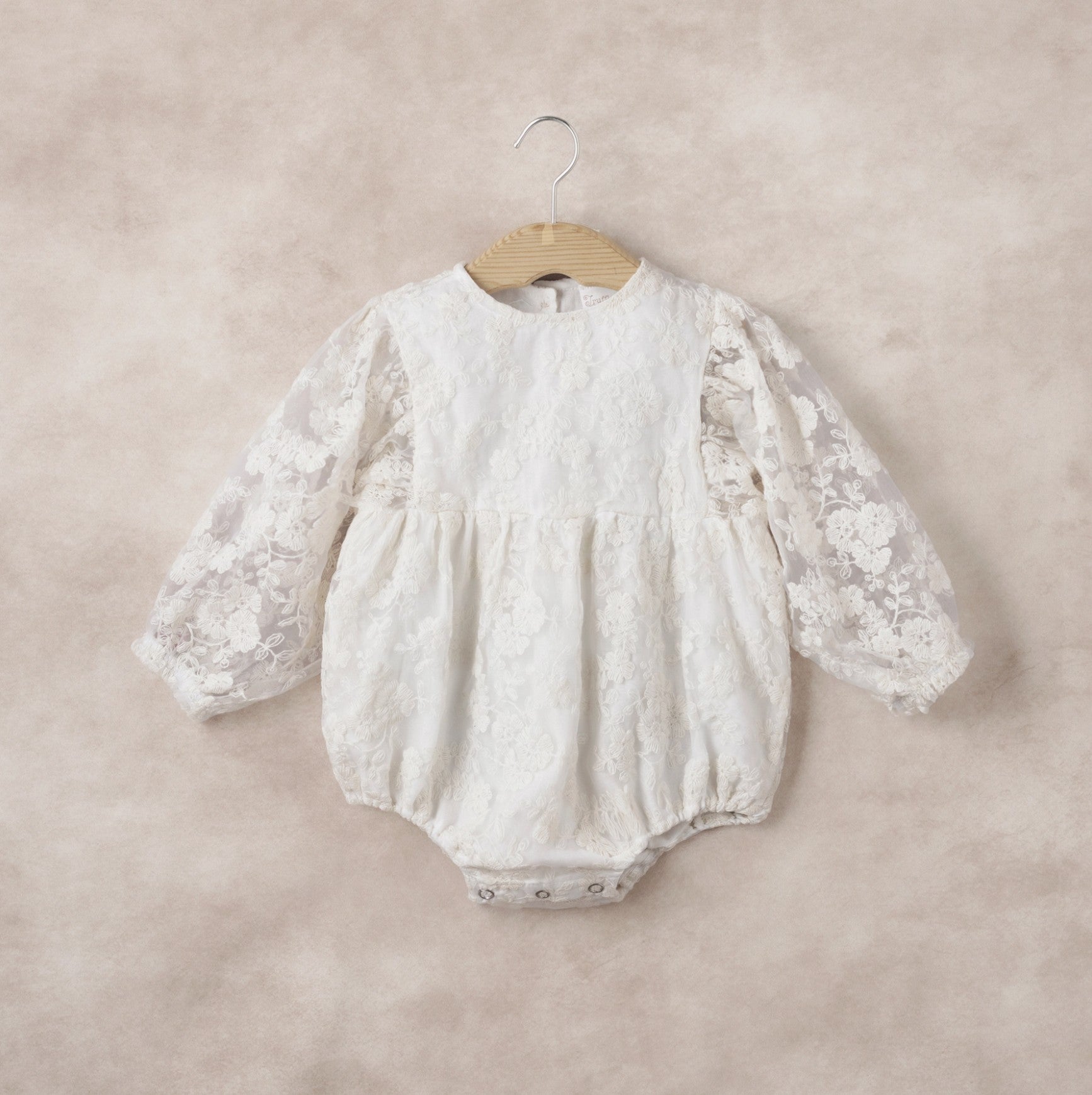 White lace baby romper on a wooden hanger against a beige background
