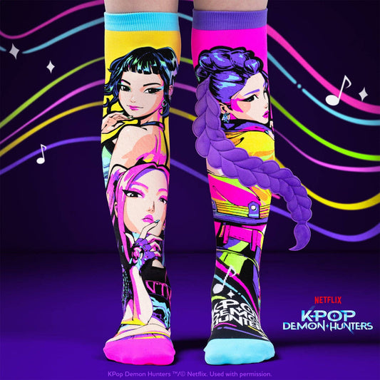 A pair of knee-high socks with vibrant, colorful K-pop character prints, shown on a stylized dark purple background with neon lines and music notes. The socks feature detailed illustrations of characters with braided hair in pink, yellow, purple, and blue.