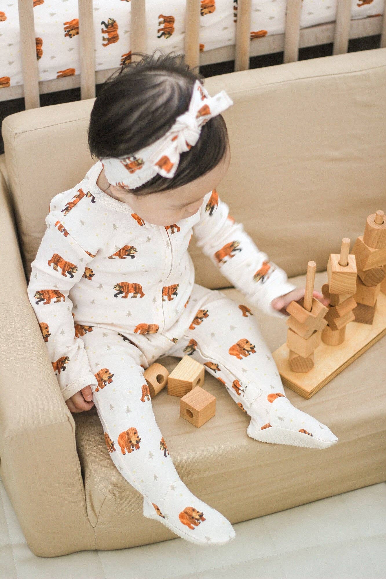 A baby sitting on a beige sofa wearing a white footed pajama set and matching headband with an orange-brown bear print, playing with wooden blocks.
