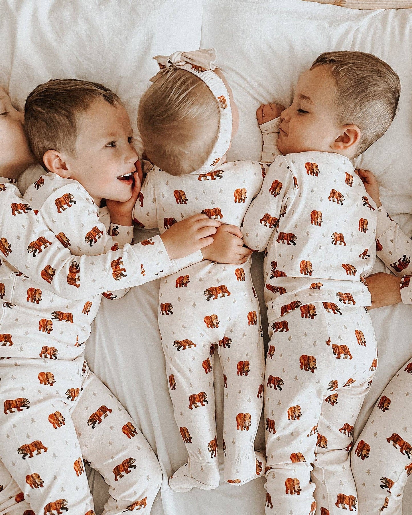 Three children lying side by side on white bedding, wearing matching cream-colored footed pajamas with a brown bear print and a small bow headband.