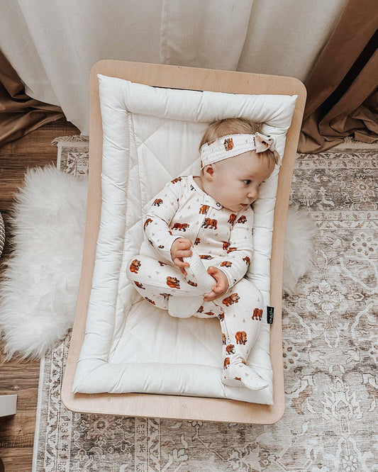 Baby in a crib with a white bedding, wearing a white outfit with brown bear patterns and a headband.
