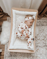 Baby in a crib with a white bedding, wearing a white outfit with brown bear patterns and a headband.
