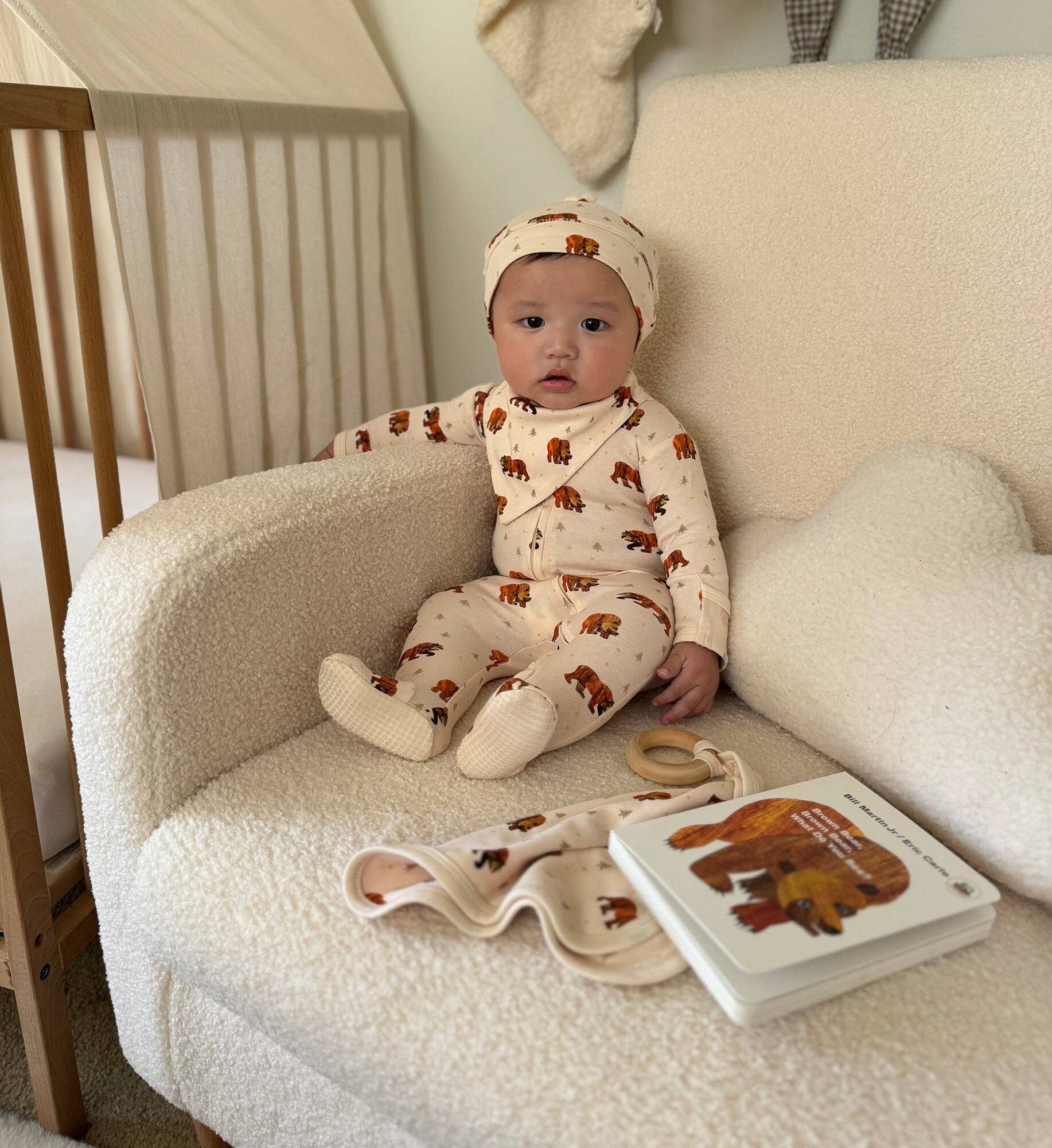 A baby wearing a cream-colored one-piece sleeper with brown bear prints and a matching beanie sits on a cream boucle armchair next to a wooden crib. A matching folded blanket and a baby book with a bear illustration are placed on the chair.