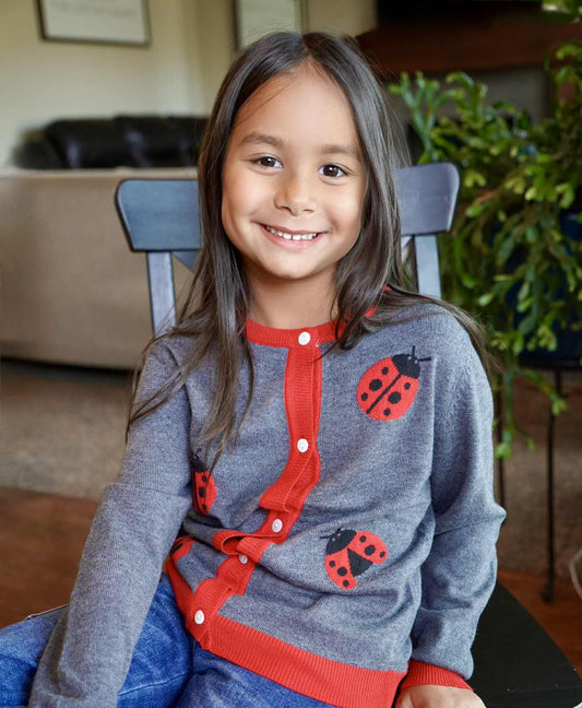 Young girl wearing a gray sweater with red accents and ladybug patterns, sitting on a chair indoors.