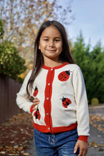 Child wearing a white cardigan with red ladybug patterns outdoors.