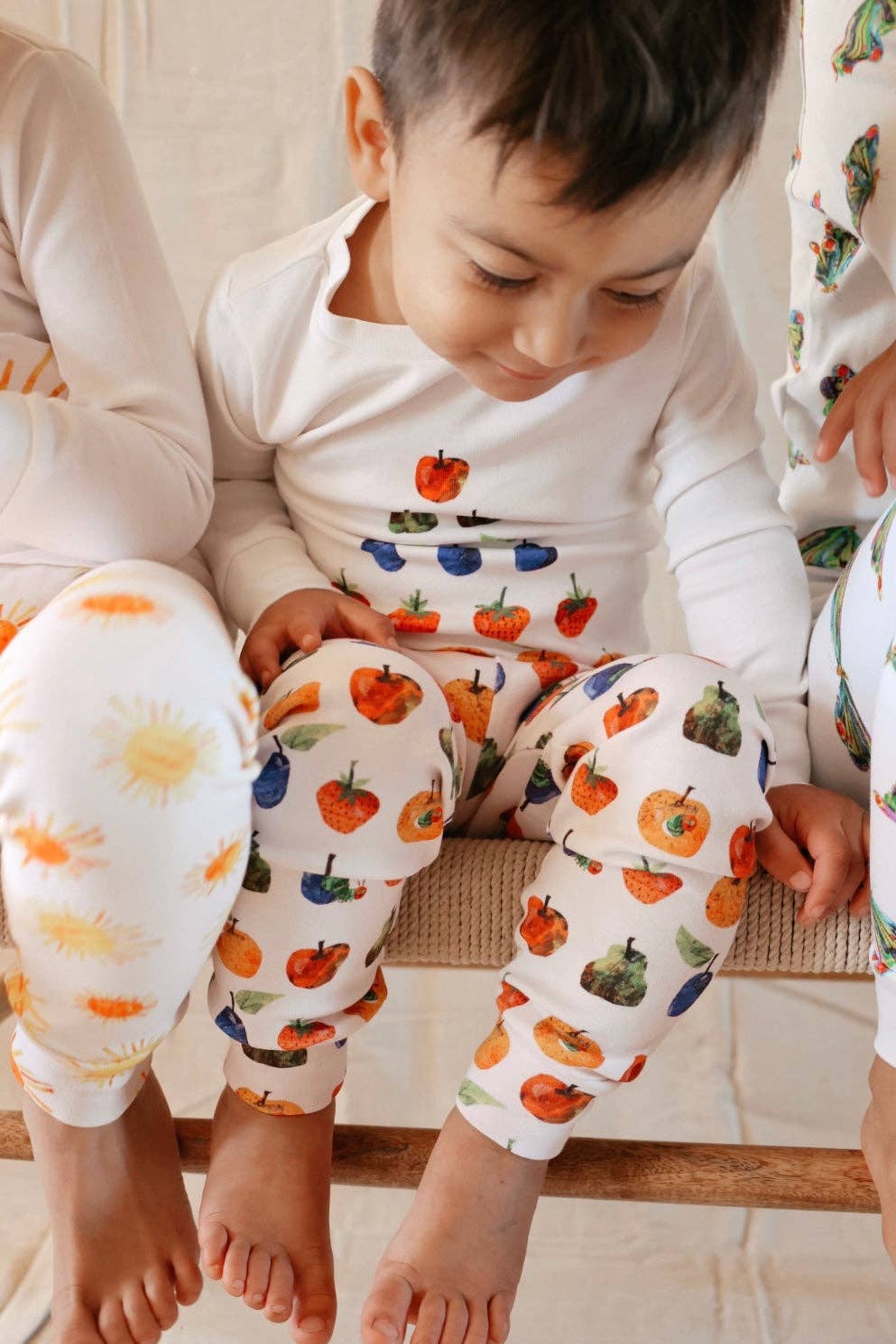 A child wearing a white long-sleeve pajama top and matching pants with a colorful fruit print, sitting on a woven bench with other children partially visible.