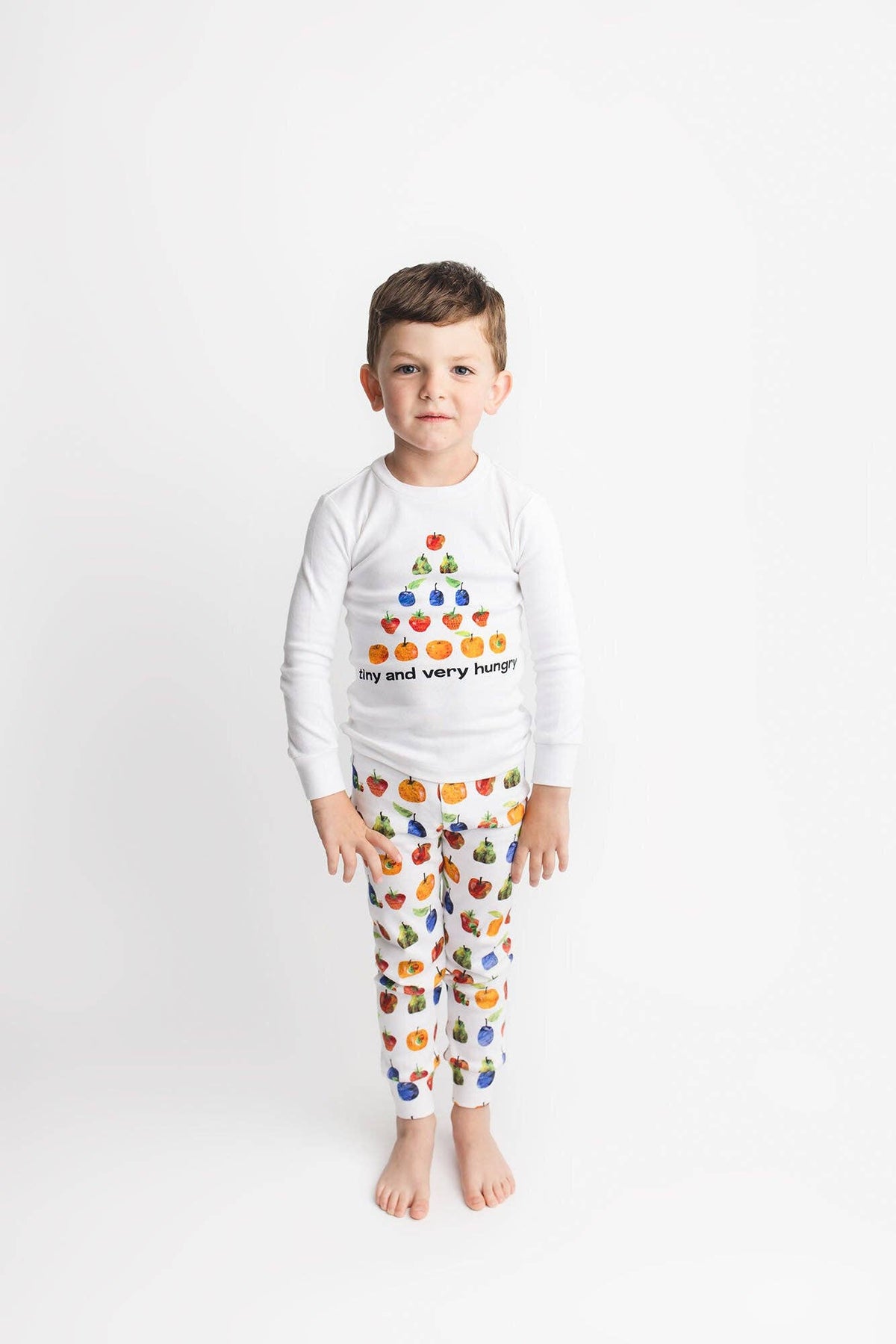 A young boy stands against a plain white studio background wearing a white long-sleeve pajama top with a colorful fruit graphic and the text 'my and very hungry' paired with matching patterned pajama pants.