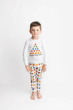 A young boy stands against a plain white studio background wearing a white long-sleeve pajama top with a colorful fruit graphic and the text 'my and very hungry' paired with matching patterned pajama pants.