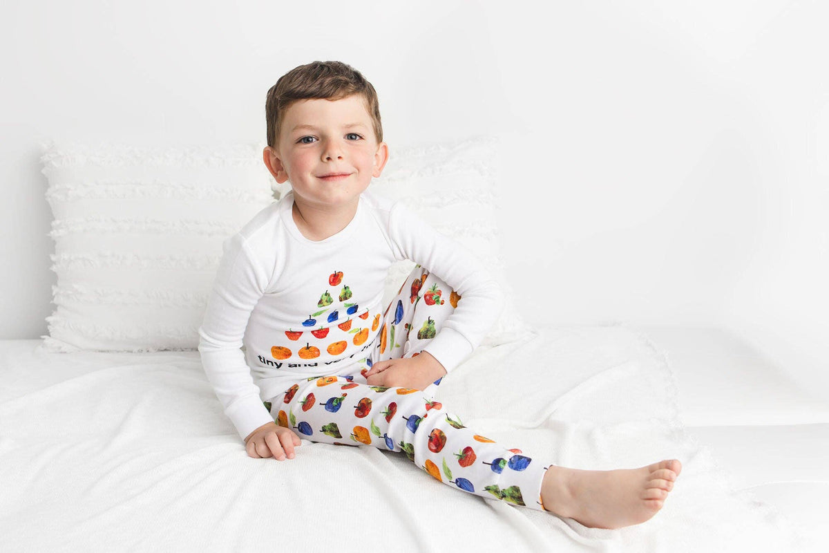 A young boy sitting on a white bed wearing a white long-sleeve pajama top and matching pajama pants with colorful fruit prints, posed against a clean white background.