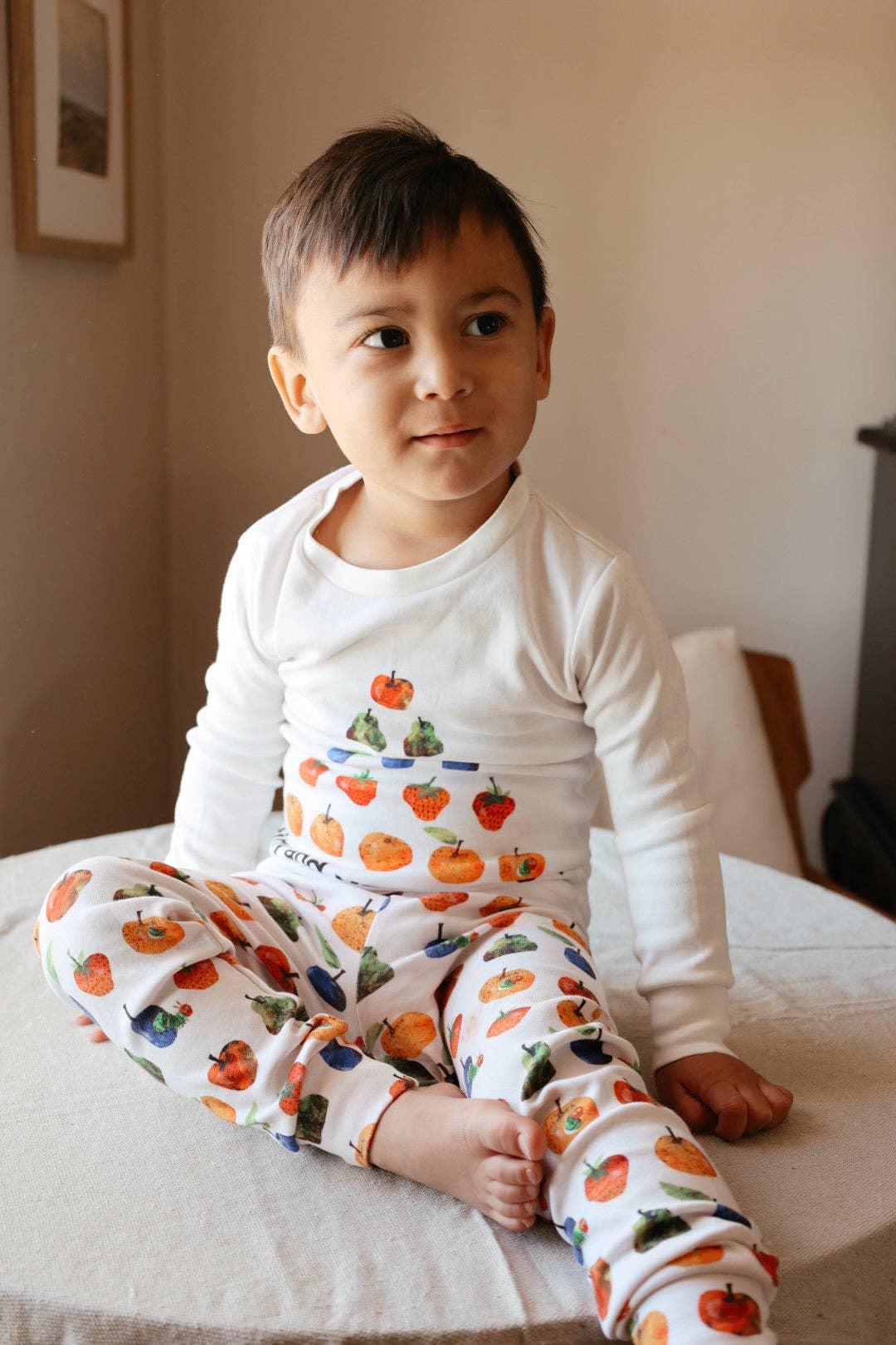 A young child wearing a white long-sleeve pajama top and matching pants with a colorful fruit print, sitting on a bed in a softly lit room.