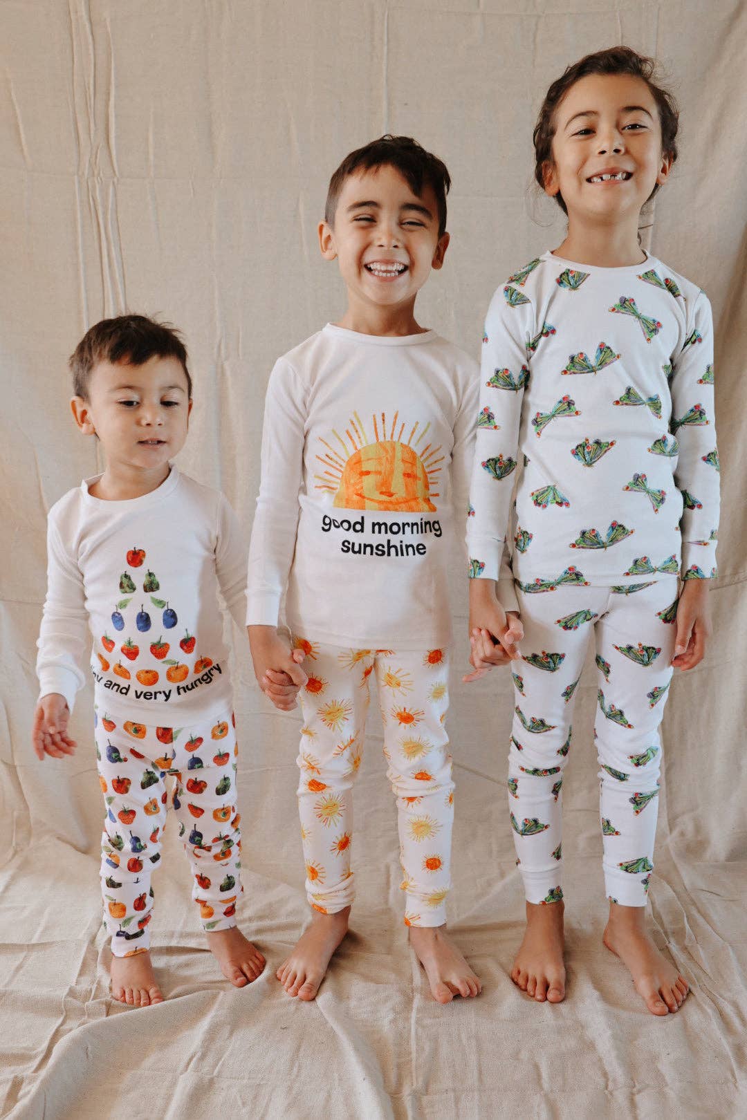 Three children standing barefoot in front of a beige fabric backdrop, wearing matching white long-sleeve pajamas with colorful printed graphics. The center child's pajama top has a sun graphic and the text 'good morning sunshine,' while the other two pajama sets feature different fruit and vegetable prints.