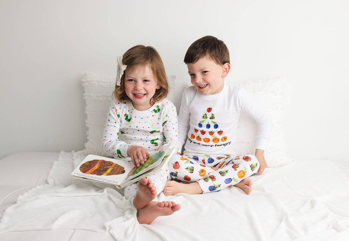 Two children sitting on a white bed reading a book. The girl wears a white long-sleeve pajama top with green cartoon characters and matching patterned pants.