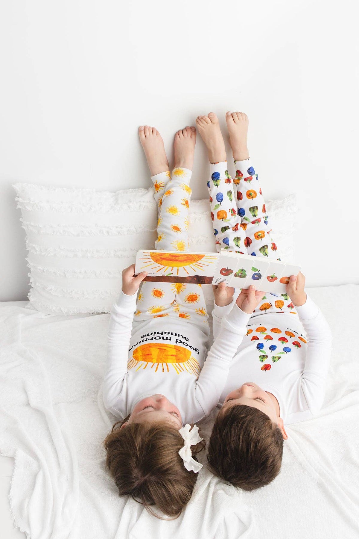 Two children lying on a white bed reading a book, wearing matching white long-sleeve pajamas with colorful sun and fruit prints.