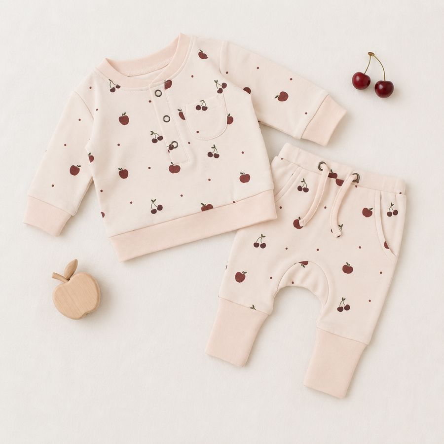A baby two-piece outfit set consisting of a long-sleeve sweatshirt and matching pants, laid flat on a light cream background. The outfit is light pink with a pattern of small red cherries and tiny red dots, finished with solid light pink ribbed cuffs and hem.