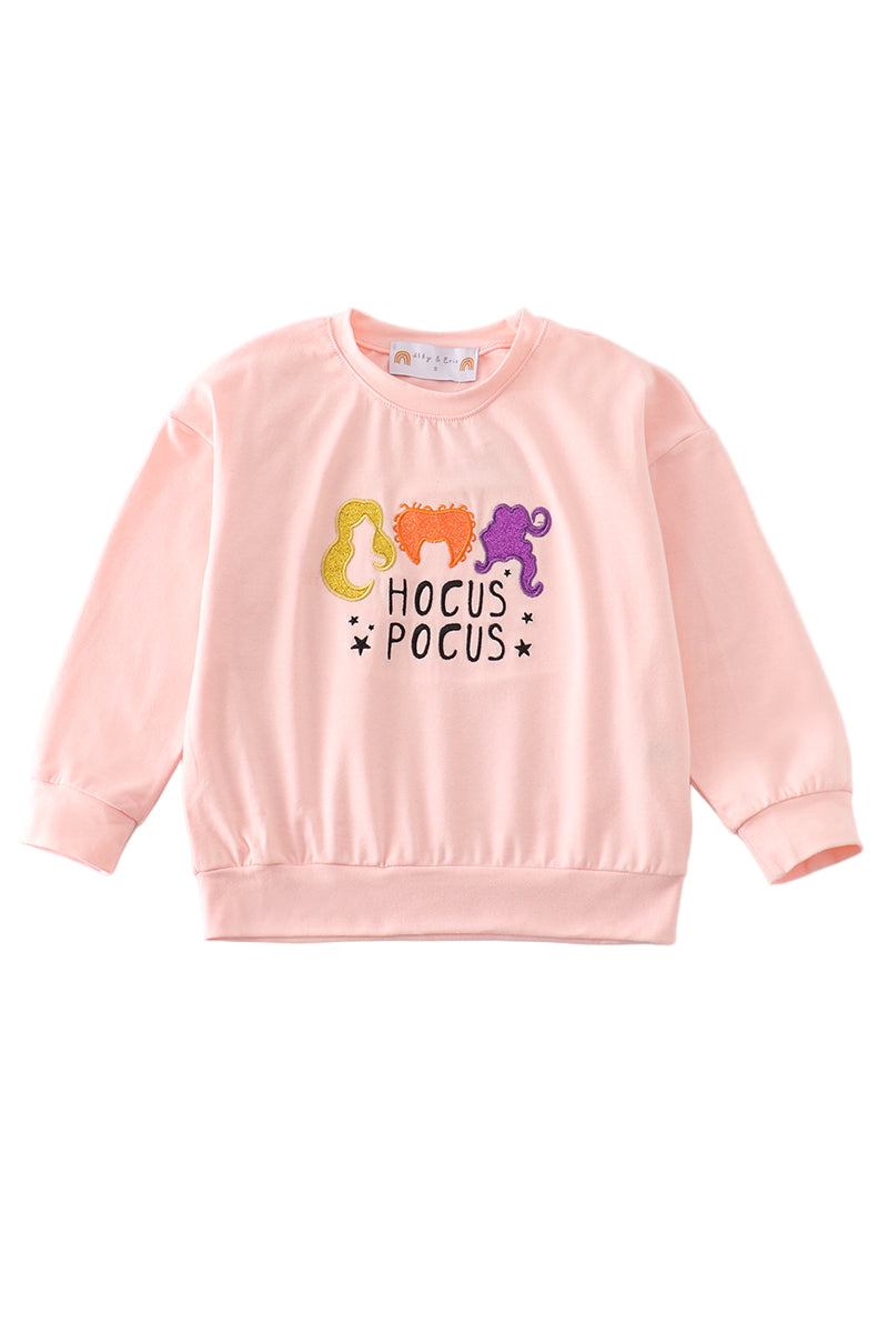 A pink long sleeve top with 'HOCUS POCUS' text and glittery decorations in the shape of witches.