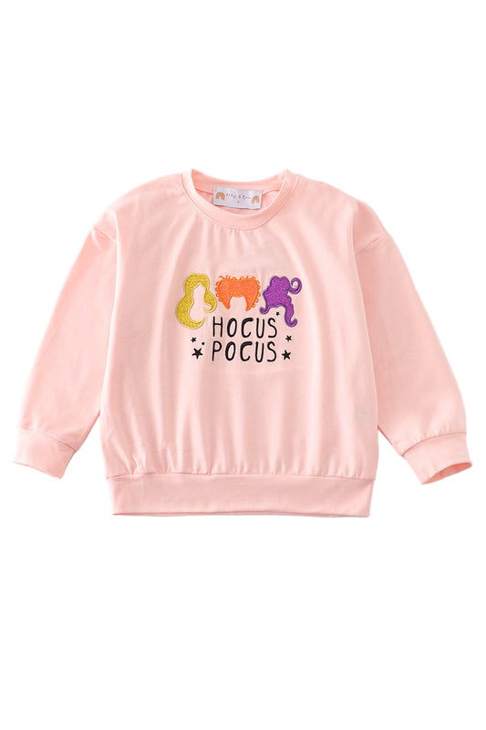 A pink long sleeve top with 'HOCUS POCUS' text and glittery decorations in the shape of witches.