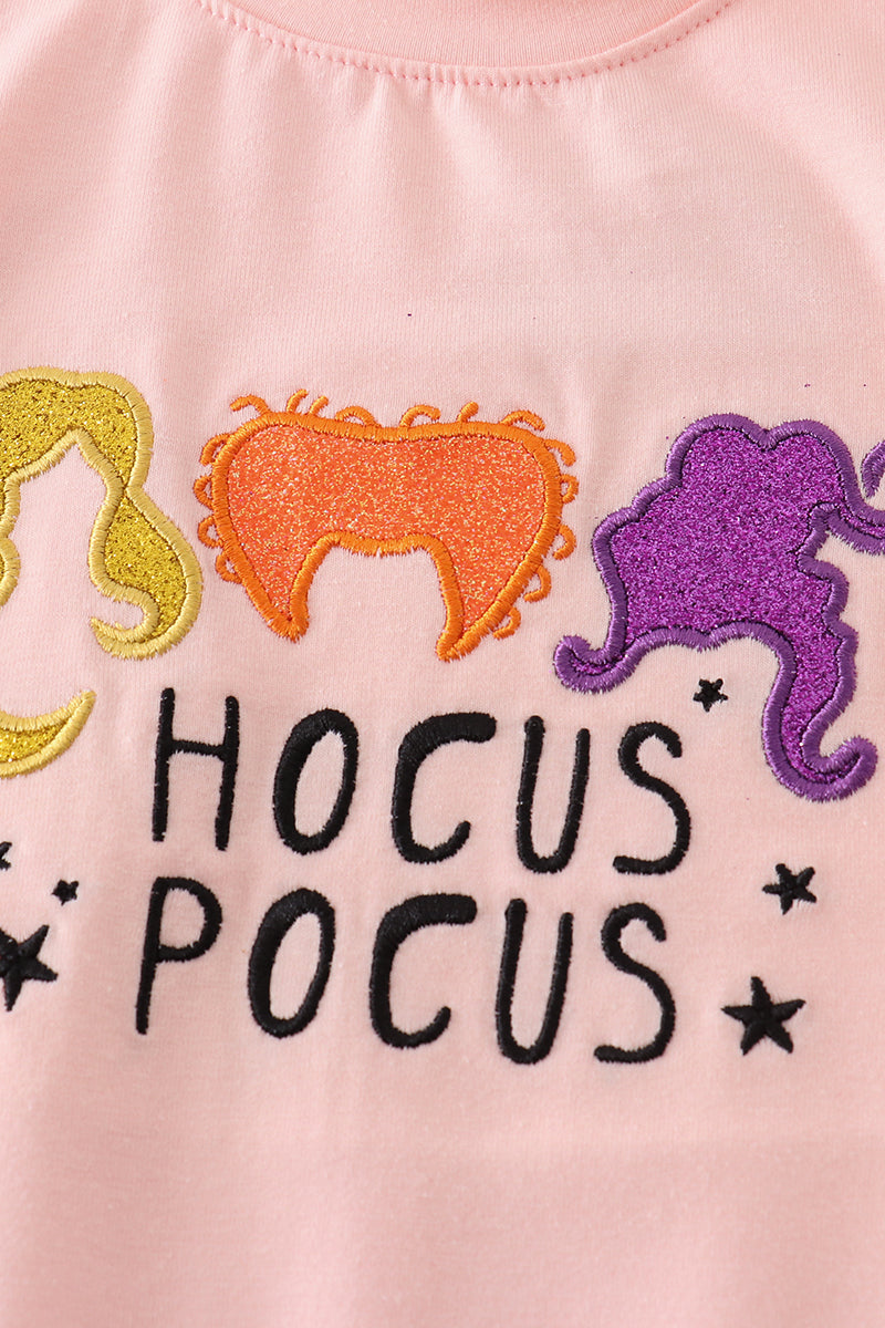 Pink shirt with 'HOCUS POCUS' text and colorful embroidered designs on a plain background