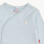 A close-up of a light blue baby wrap sleeper with a small white embroidered bird on the chest. The inner neck label shows the brand 'magnetic me' and a 'eco-friendly' tag.