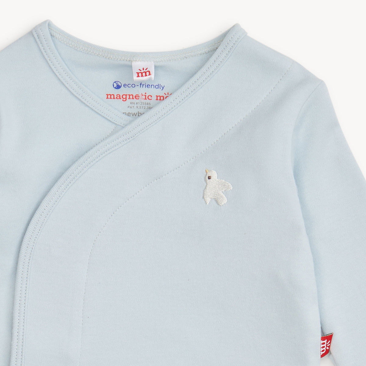 A close-up of a light blue baby wrap sleeper with a small white embroidered bird on the chest. The inner neck label shows the brand 'magnetic me' and a 'eco-friendly' tag.
