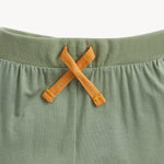 Close-up of a sage green knit garment, likely a baby romper or shorts, featuring a gathered waistband and a contrasting mustard yellow braided drawstring tied in an X.
