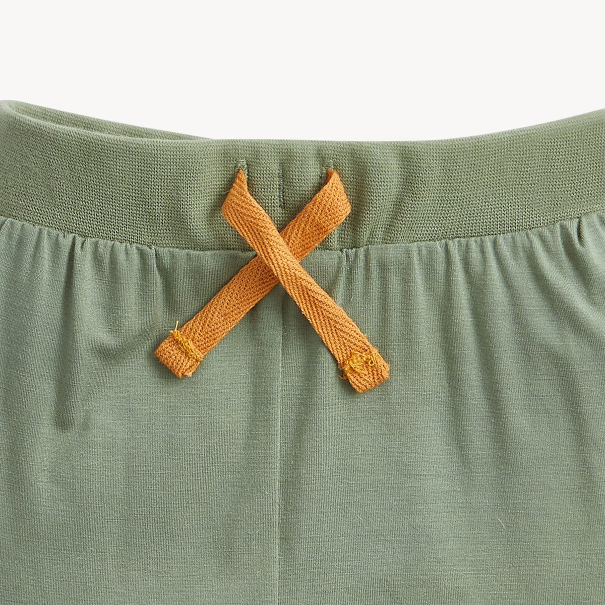 Close-up of a sage green knit garment, likely a baby romper or shorts, featuring a gathered waistband and a contrasting mustard yellow braided drawstring tied in an X.