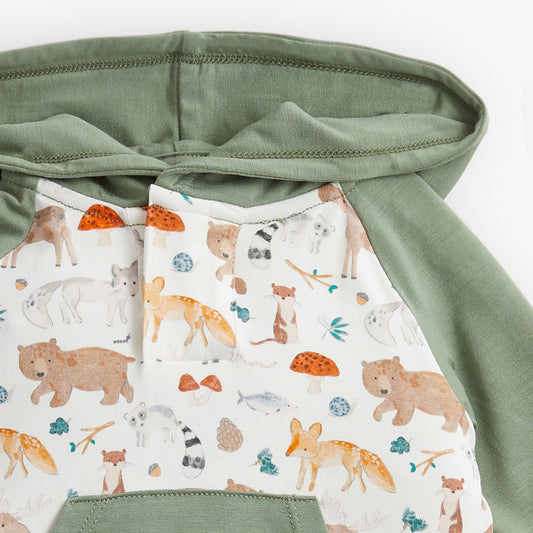 Close-up of a children's hooded garment with sage green fabric on the hood and sleeves, and a white front panel printed with woodland animals including foxes, bears, a raccoon, mushrooms, and small blue and orange accents.