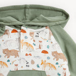 Close-up of a children's hooded garment with sage green fabric on the hood and sleeves, and a white front panel printed with woodland animals including foxes, bears, a raccoon, mushrooms, and small blue and orange accents.