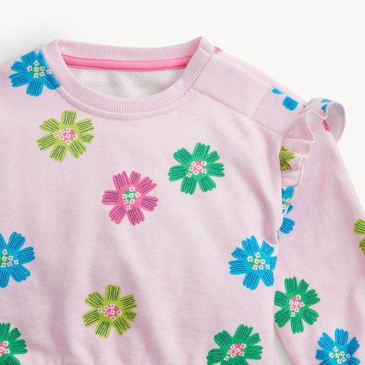 A close-up of a light pink long-sleeve top with colorful embroidered flowers in blue, green, and pink, featuring a ruffled sleeve detail.