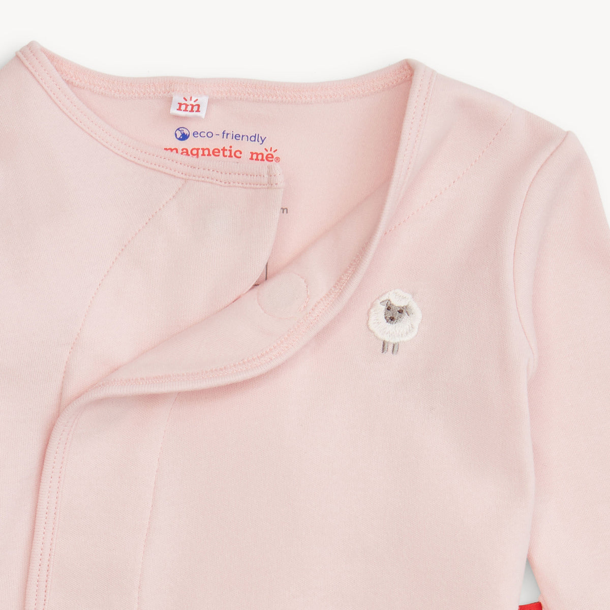 Close-up of a light pink long-sleeve baby wrap or sleeper with a snap closure and a small sheep applique on the chest. The inside neck label shows the brand 'mii' and text reading 'eco-friendly magnetic me.