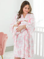 Woman in a toile robe holding a baby in a room with a pink chair and white crib.