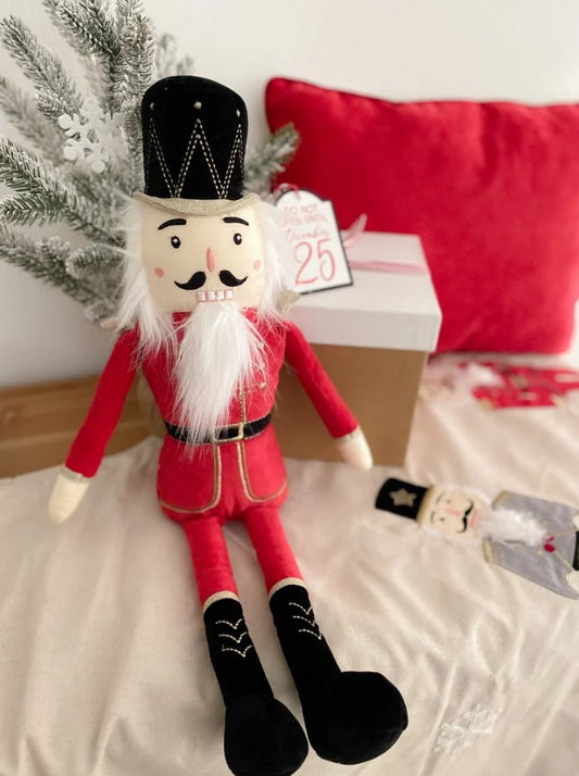 A plush nutcracker doll dressed in a red coat and black hat, posed on a bed with a red pillow, gift box, and small tag that reads 'Merry 25th'.