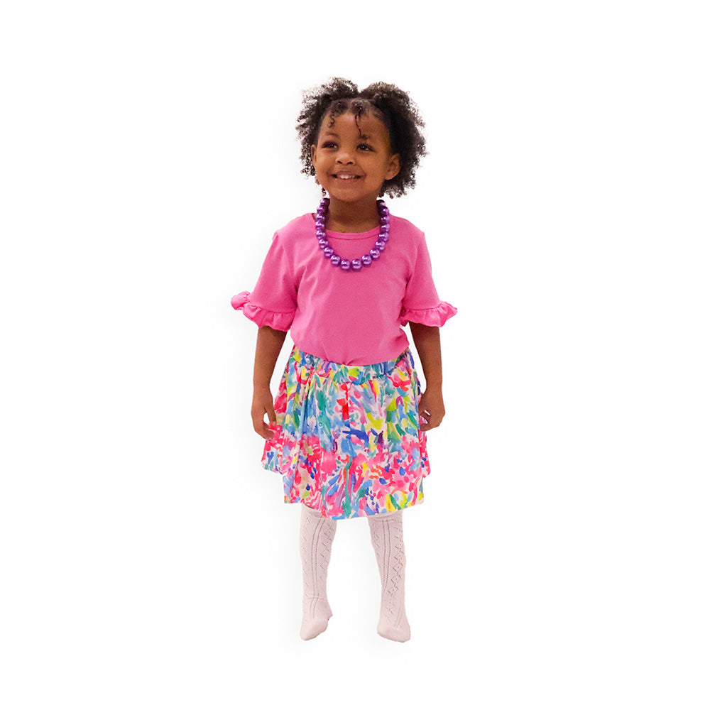 A child model wearing a pink floral print skort set with a matching pink top and white tights.