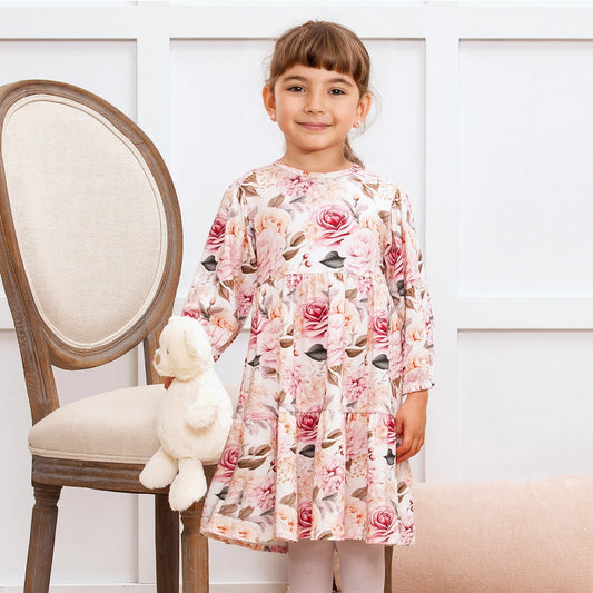 Young girl in a floral dress holding a teddy bear, standing next to a wooden chair with a white cushion.