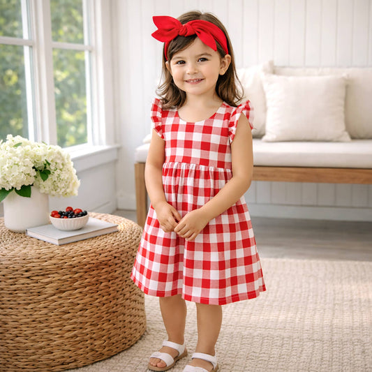 Young girl in a red and white checkered dress with a red bow standing in a bright room.