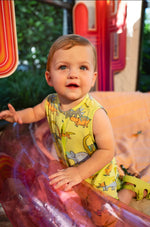 Baby wearing a yellow sleeveless romper with dinosaur prints, sitting on a colorful surface.