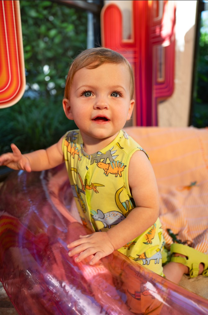Baby wearing a yellow sleeveless romper with dinosaur prints, sitting on a colorful surface.